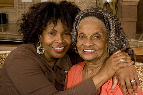 Young woman with arms around the shoulders of senior woman sitting on couch smiling